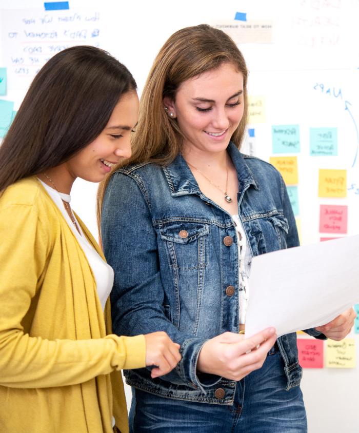 A photo of two women in front of a white board reading a document together.